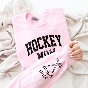 May include: Pink sweatshirt with the text "HOCKEY MOM" in black on the front and the text "JOHN SOPHIA" with a hockey stick and puck graphic on the sleeve.