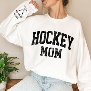 May include: White sweatshirt with black text that reads "HOCKEY MOM" in a vintage style font. The sweatshirt has a crew neck and long sleeves.