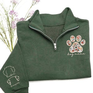 May include: Dark green quarter-zip sweatshirt with a floral paw print design and the words "dog mama." The sleeve features embroidered dog silhouettes with the names "Tucker" and "Hooper." The sweatshirt has a silver zipper and a high collar.