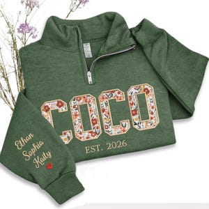 May include: Green quarter-zip sweatshirt with the name "COCO" embroidered in floral letters. The text "EST. 2026" is below the name. The left sleeve has the names "Ethan, Sophia, Kaity" embroidered, with a red heart.