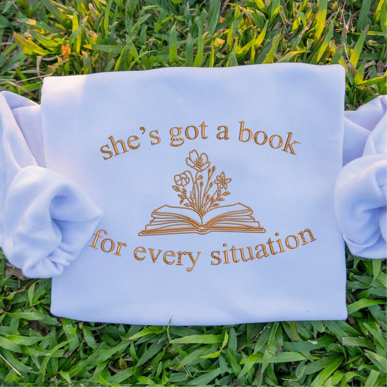 May include: A light blue sweatshirt with the text "she's got a book for every situation" in gold embroidery. The text is centred on the sweatshirt and is surrounded by a gold embroidered illustration of a book with flowers growing from it.