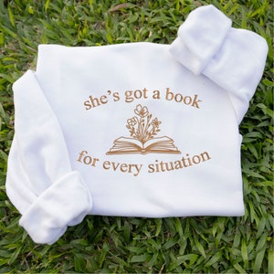 May include: White baby onesie with brown embroidery that reads "she's got a book for every situation" with a stylised image of a book with flowers on top.
