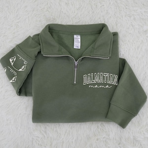 May include: Olive green pullover sweatshirt with a quarter-zip neckline. The front features the text "DALMATIAN mama" in white script. The left sleeve has Dalmatian dog illustrations and the names "Charlie" and "Willie" in white.