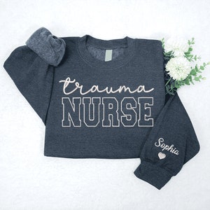 May include: A dark gray sweatshirt with the text "trauma NURSE" in white with a black outline. The text "Sophia" is embroidered on the sleeve in pink with a small heart.