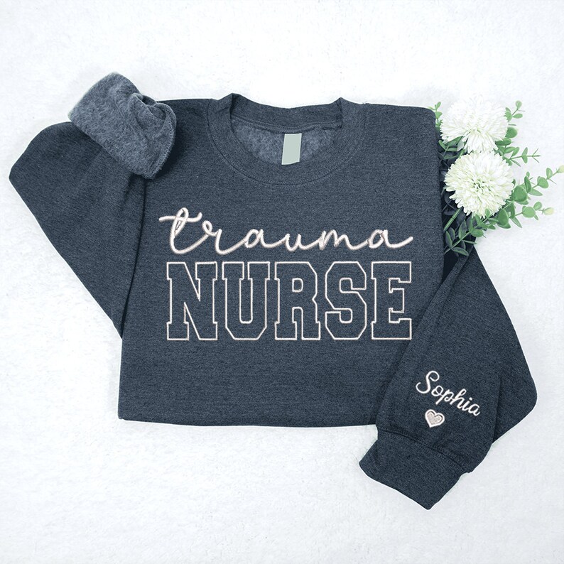 May include: A dark gray sweatshirt with the text "trauma NURSE" in white with a black outline. The text "Sophia" is embroidered on the sleeve in pink with a small heart.