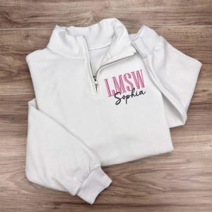 May include: White quarter-zip sweatshirt with long sleeves. The sweatshirt features a silver zipper and the text "LMSW Sophia" in pink and black script on the front. The garment is folded on a wooden surface.