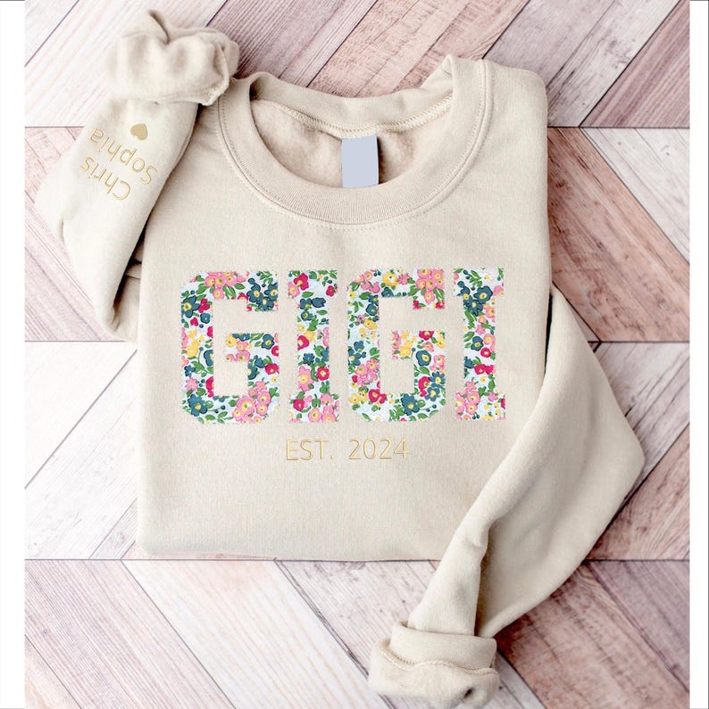 May include: A beige sweatshirt with a floral print design that spells out "GIGI" and the text "EST. 2024" in gold lettering.