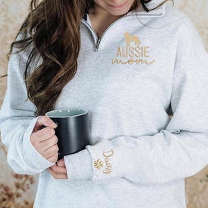 May include: A light gray sweatshirt with a quarter-zip neckline. The sweatshirt features gold embroidery of an Australian Shepherd dog, the words "AUSSIE mom", the name "Daisy", and a paw print. A black mug is held in the hands.