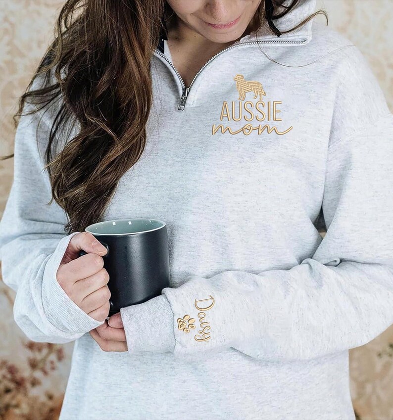 May include: A light gray sweatshirt with a quarter-zip neckline. The sweatshirt features gold embroidery of an Australian Shepherd dog, the words "AUSSIE mom", the name "Daisy", and a paw print. A black mug is held in the hands.