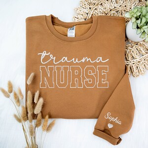 May include: A brown sweatshirt with the text "trauma NURSE" in white embroidery. The text "Sophia" is embroidered on the sleeve with a small heart.