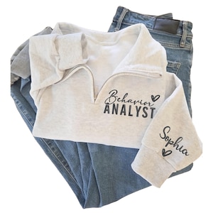 May include: A light gray sweatshirt with a quarter-zip neckline, the words "Behavior Analyst" are printed in black script on the front. The sleeve has the name "Sophia" printed in black script. The sweatshirt is paired with blue denim jeans.