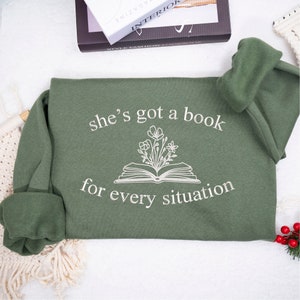 May include: A green sweatshirt with the text "she's got a book for every situation" and a drawing of a book with flowers growing out of it.