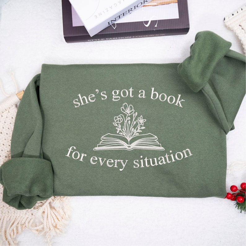 May include: A green sweatshirt with the text "she's got a book for every situation" and a drawing of a book with flowers growing out of it.