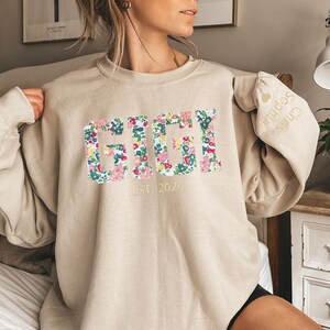 May include: A beige sweatshirt with a floral print that says "GIGI EST. 2024" on the front. The sleeve has a small embroidered heart with the text "Chris Sophia" on it.