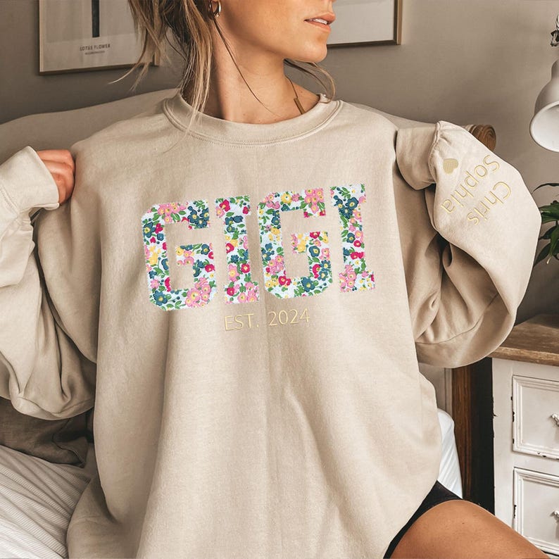 May include: A beige sweatshirt with a floral print that says "GIGI EST. 2024" on the front. The sleeve has a small embroidered heart with the text "Chris Sophia" on it.