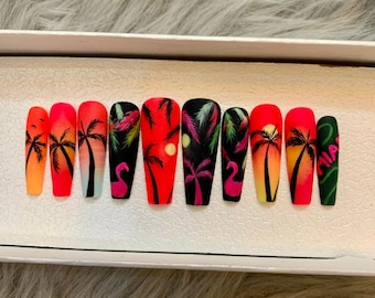May include: Set of ten false nails with a tropical sunset design. The nails feature palm trees, flamingos, and a sunset gradient in shades of orange, pink, and yellow. The nails are also decorated with black and green accents.