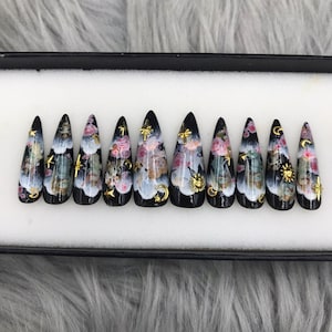 Könnte beinhalten: Ein Set mit zehn schwarzen Stiletto-Kunstnägeln mit einem floralen und himmlischen Design. Die Nägel haben goldene Akzente, darunter Sterne, Monde und eine Sonne.
