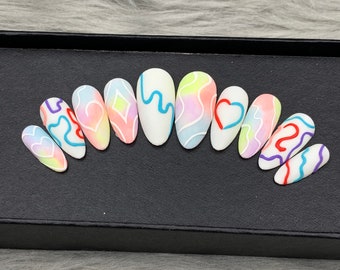Puede incluir: Un juego de diez uñas postizas en forma de almendra con una base blanca mate y diseños pintados a mano. Los patrones incluyen lavados de acuarela en tonos pastel, líneas onduladas en azul, rojo y morado, y formas de corazón y diamante.