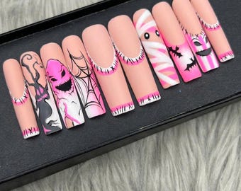 May include: A set of long, square-shaped false nails with a Halloween theme. Each nail features a different design, including a mummy, bats, a jack-o'-lantern, and a spiderweb, all in pink, black, and white. The nails are displayed on a black surface.