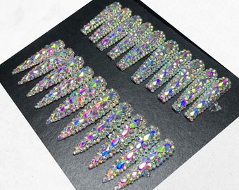 Può includere: Un set di 10 ciondoli per unghie iridescenti, ogni ciondolo ha la forma di un chiodo lungo, sottile e appuntito con una fila di piccoli strass iridescenti.
