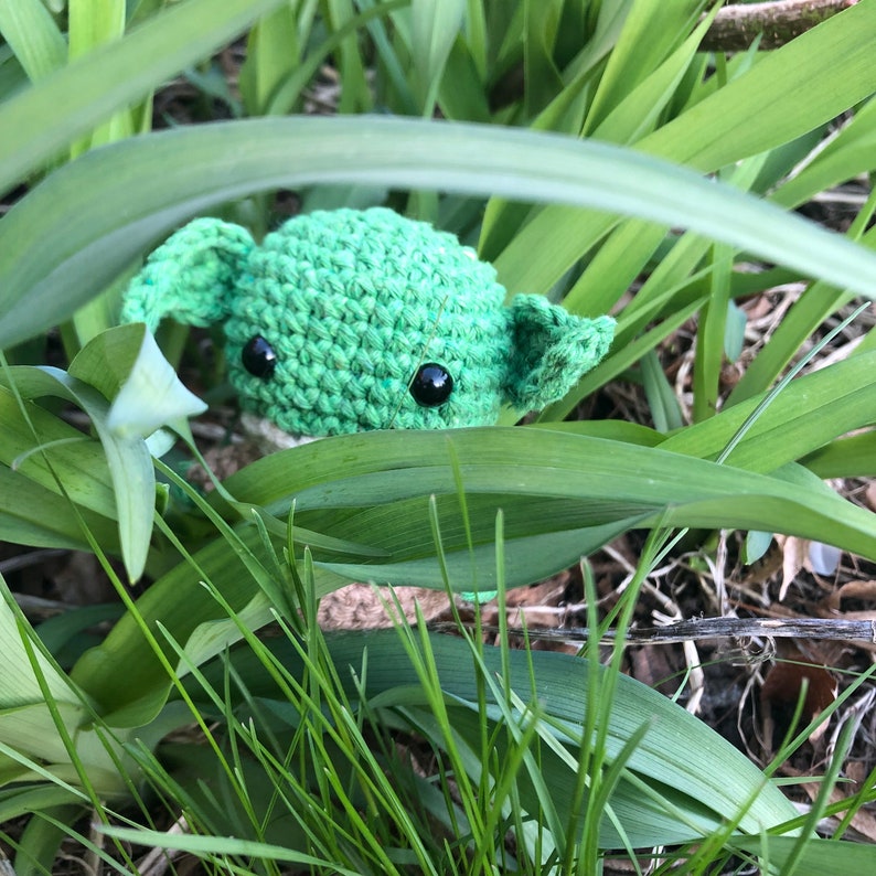 May include: A green crocheted Yoda character with black eyes peeking out from behind tall green blades of grass.
