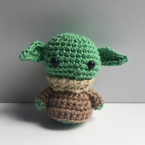 May include: A crocheted green and brown plush toy resembling a popular fictional character. The toy has large black eyes and small ears.