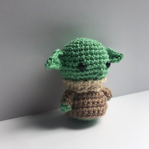 May include: A green crocheted plush toy with large black eyes and a brown crocheted sweater. The toy is a popular character from the Star Wars franchise.