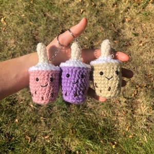 May include: Three crocheted bubble tea keychains in pink, purple, and beige. Each plush cup features a white whipped cream top, a protruding straw, and a stitched smiling face with black bead eyes. The keychains are attached to metal rings.