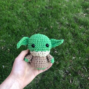May include: A green and brown crocheted plush toy resembling a character from the Star Wars franchise. The toy has large eyes and a small mouth.