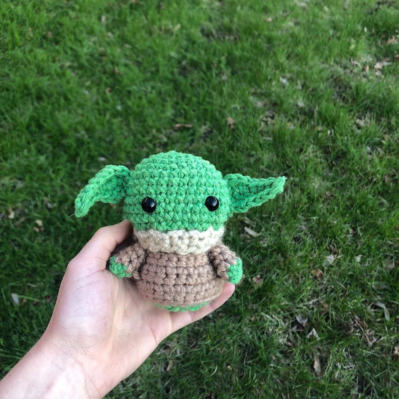 May include: A green and brown crocheted plush toy resembling a character from the Star Wars franchise. The toy has large eyes and a small mouth.