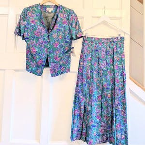 May include: A vintage two-piece outfit comprising a short-sleeved jacket and a pleated skirt. The set is teal green with a floral pattern in purple, pink, and blue. The jacket has a V-neck and button closure. The skirt is long and flowing.