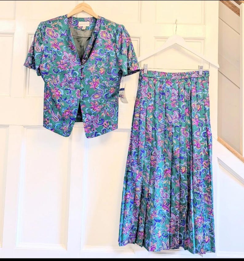 May include: A vintage two-piece outfit comprising a short-sleeved jacket and a pleated skirt. The set is teal green with a floral pattern in purple, pink, and blue. The jacket has a V-neck and button closure. The skirt is long and flowing.