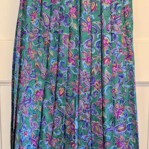 May include: A long, pleated skirt with a vibrant floral pattern. The skirt has a green background with purple, blue, and yellow floral and paisley designs. The skirt is hanging on a metal hanger.