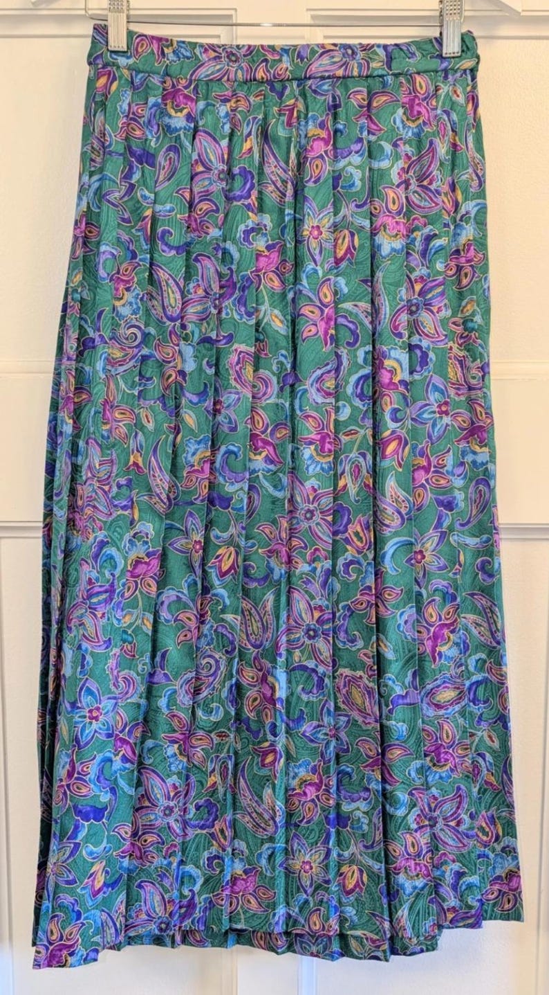 May include: A long, pleated skirt with a vibrant floral pattern. The skirt has a green background with purple, blue, and yellow floral and paisley designs. The skirt is hanging on a metal hanger.
