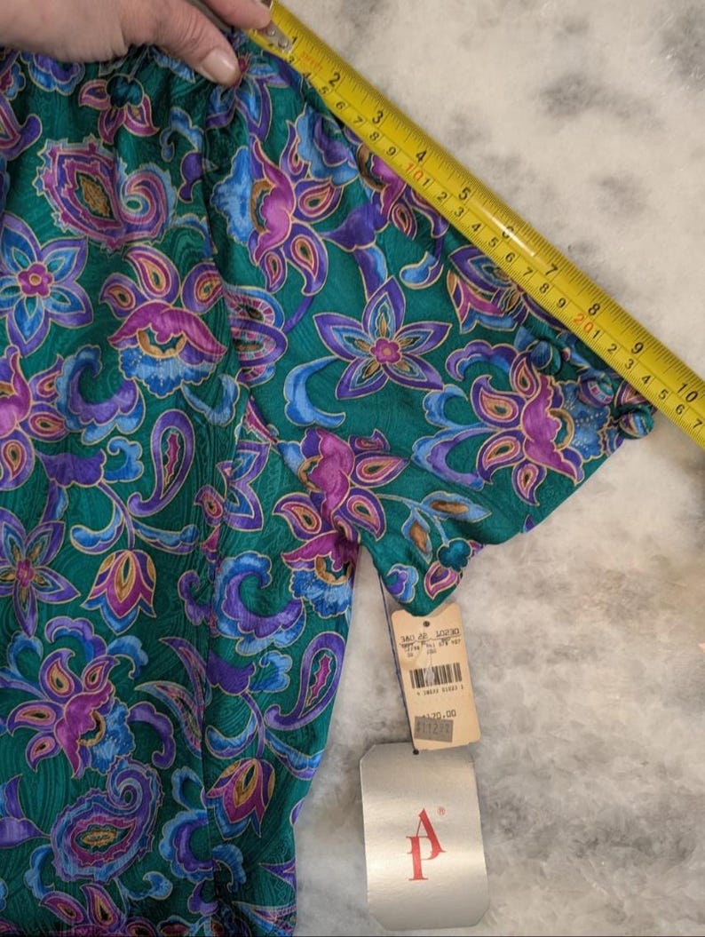 May include: A short-sleeved top with a vibrant floral and paisley pattern in shades of teal, purple, and blue. The fabric is measured with a yellow tape measure. A price tag is attached. The top has a gathered neckline and sleeves.