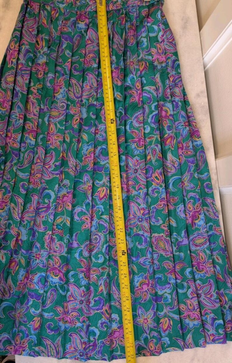 May include: A long, pleated skirt with a vibrant floral pattern in shades of green, purple, and blue. A yellow measuring tape shows the skirt's length. The skirt's length is approximately 76 cm.