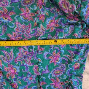 May include: A green garment featuring a colourful floral and paisley design in purple, pink, and blue. A yellow tape measure is used to measure the fabric. The garment has a button closure.
