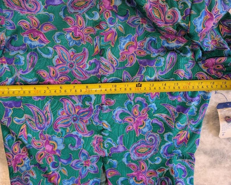 May include: A green garment featuring a colourful floral and paisley design in purple, pink, and blue. A yellow tape measure is used to measure the fabric. The garment has a button closure.