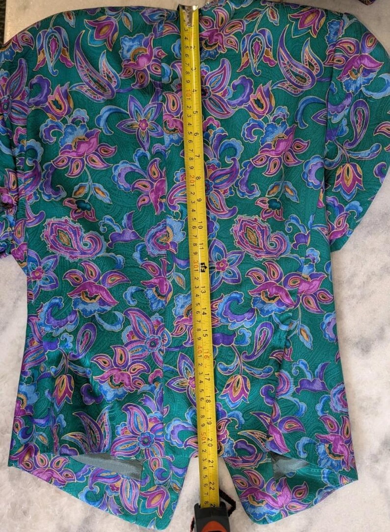 May include: A green blazer featuring a floral design in purple, pink, and blue. A yellow tape measure shows the blazer's length to be approximately 56 cm.