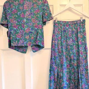 May include: A vintage two-piece outfit comprising a short-sleeved jacket and a pleated skirt. The set displays a colourful floral pattern in green, purple, pink, and blue. The jacket is buttoned, and the skirt is pleated.