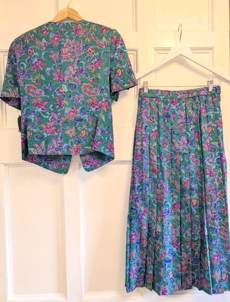 May include: A vintage two-piece outfit comprising a short-sleeved jacket and a pleated skirt. The set displays a colourful floral pattern in green, purple, pink, and blue. The jacket is buttoned, and the skirt is pleated.