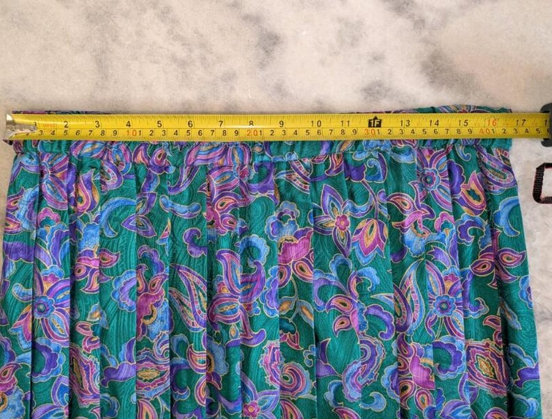 May include: A green skirt with a floral paisley pattern in shades of purple, blue, and pink. The skirt measures approximately 43 cm across the top, as measured by a yellow tape measure.