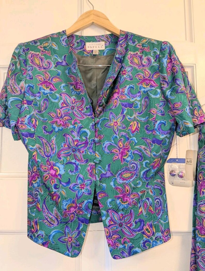 May include: A vintage green two-piece suit with a floral paisley pattern in purple, blue, and yellow. The jacket has short sleeves and a button-up front. The label reads "Papell".