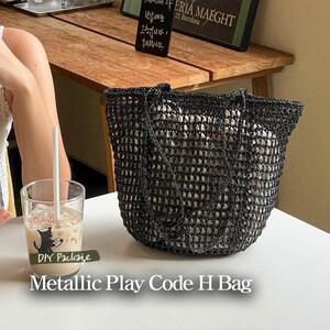 May include: A black mesh tote bag with a metallic sheen. The bag has a wide, open top and two long handles. The bag is sitting on a table with a glass of iced coffee in front of it.