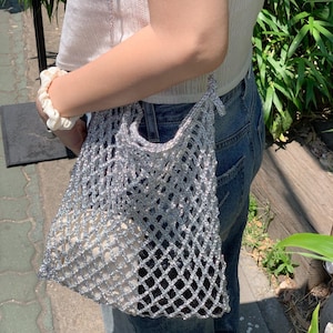 May include: A silver mesh tote bag with a white lining. The bag is being carried by a person wearing a white top and blue jeans.