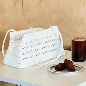 May include: White crocheted tote bag with a wide, flat base and two handles. The bag is sitting on a white table with a small plate of two brown pastries and a glass of iced coffee.