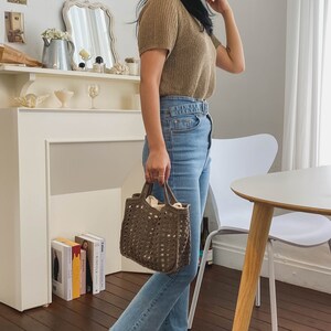 May include: A woman wearing a brown knitted sweater and blue jeans is holding a brown crocheted tote bag.
