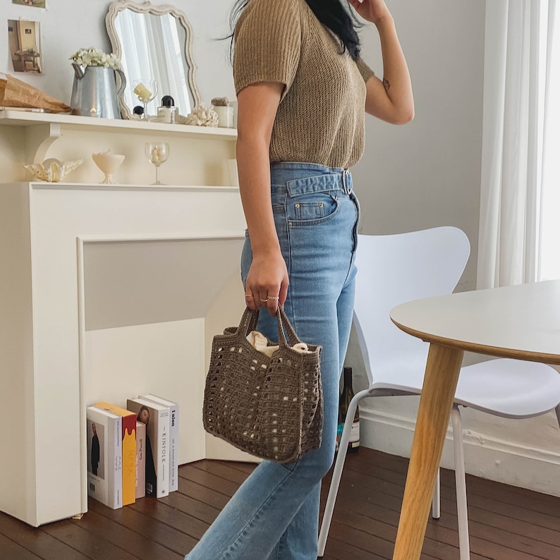 May include: A woman wearing a brown knitted sweater and blue jeans is holding a brown crocheted tote bag.
