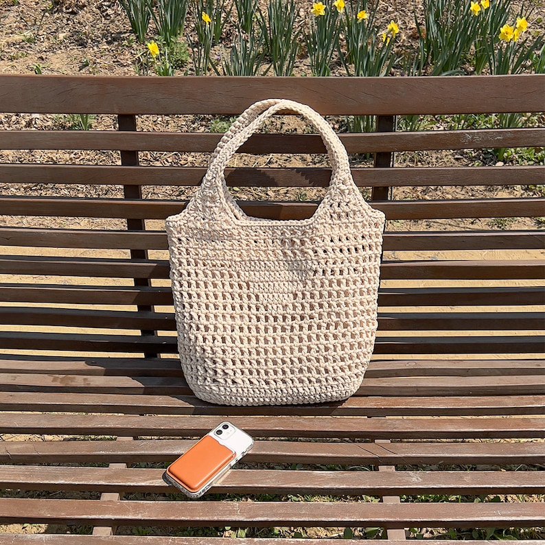 Puede incluir: Una bolsa de mano de crochet blanca con un patr&oacute;n de tejido abierto grande. La bolsa est&aacute; sentada en un banco de madera.