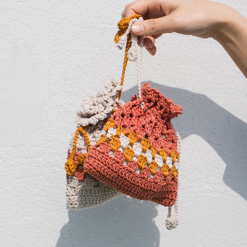 May include: Two crocheted drawstring bags, one in a beige color and the other in a red, white, and yellow color pattern. The bags are hanging from a hand.